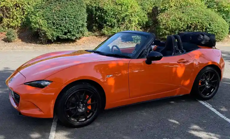 119 / 5,000Mazda MX5 orange avec toit baissé photographiée de face avec un porte-bagages d'origine Boot-bag monté sur le couvercle du coffre